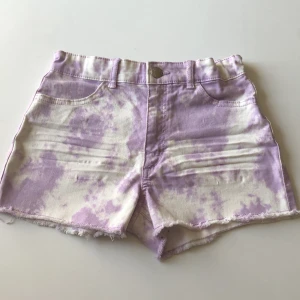 Lila och vita shorts från H&M - Snygga lila och vita shorts från H&M i tie-dye stil. De har hög midja, råa kanter och klassiska fickor både fram och bak. Perfekta för sommaren och riktigt trendiga med sin unika färgkombination. Gjorda i mjuk bomull för maximal komfort.