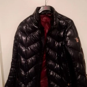 Svart dunjacka från Moncler - Säljer en svart glansig dunjacka från Moncler med klassisk logga på ärmen. Jackan har quiltad design, dragkedja framtill och röd insida. Perfekt för kalla dagar och riktigt snygg till streetwear-stilen.