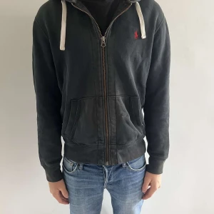 Svart hoodie från Ralph Lauren - Svart hoodie från Ralph Lauren med dragkedja framtill, vit dragsko i huvan och röd broderad logga på bröstet. Jackan har kängurufickor och ribbade muddar vid ärmslut och nederkant. Perfekt för en chill och avslappnad stil.
