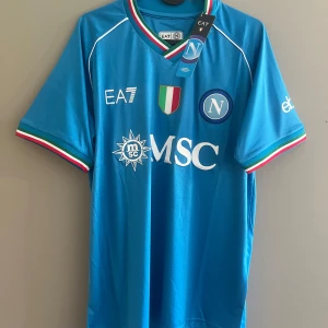 Napoli EA7 blå fotbollströja M - Napoli fotbollströja från EA7 i klarblått med vita loggor och detaljer. Trikolorfärgade muddar och krage, klubbmärke och sponsortryck på bröstet. Tillverkad i lätt och ventilerande polyester, perfekt för match eller träning.