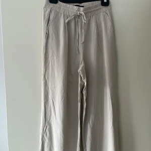 Beiga linnebyxor från Vero Moda - Snygga och luftiga beiga linnebyxor från Vero Moda. Byxorna har resår och snörning i midjan, raka vida ben och fickor både fram och bak. Fickorna därbak är stängda och går ej att öppna. Dessa linnebyxor perfekta för en chill och avslappnad stil under varmare dagar.  Dessa linnebyxor är knappt andvända och i ett mycket bra skick. Kontakta för fler bilder eller frågor.