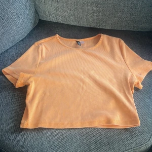 Croppad ribbad topp från H&M orange - Säljer en croppad ribbad topp från H&M i en snygg ljus orange färg. T-shirten har rund halsringning och korta ärmar, perfekt för varma dagar. Enkel och stilren design som är lätt att matcha med jeans eller kjol.Och är helt ny och är i storlek L.