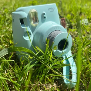 Kamera - En mintgrön Fujifilm Instax Mini 9 direktkamera, perfekt för att ta roliga och kreativa polaroidbilder med vänner. Kameran är kompakt, har en praktisk handledsrem och är enkel att använda – perfekt för spontana fotostunder och minnesvärda ögonblick.
