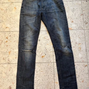 Blå jeans från The Blue Uniform - Snygga blå jeans från The Blue Uniform med slitningar och hål på knäna för en cool, avslappnad look. Klassisk femficksmodell med dragkedja och läderpatch bak i midjan. Jeansen har en rak passform och är tillverkade i bomull.