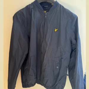 Mörkblå bomberjacka från Lyle & Scott - Snygg mörkblå bomberjacka från Lyle & Scott med klassisk gul logga på bröstet. Jackan har dragkedja framtill, ribbade muddar och två fickor med knappstängning. Perfekt för dig som gillar stilren och sportig look.