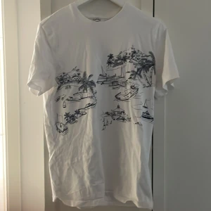 Vit t-shirt med marinblå print - Vit t-shirt med marinblå tecknade motiv av båtar, palmer och kustmiljö. Klassisk rund hals och korta ärmar. Perfekt för en avslappnad och somrig look. Tillverkad i mjuk bomull som känns skön mot huden.