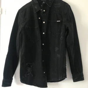 Svart jeansovershirt från Zara - Svart jeansovershirt från Zara med relaxed fit. Skjortan har slitna detaljer, tryckknappar framtill och två bröstfickor. Perfekt för en cool och avslappnad look. Långärmad och tillverkad i kraftigt jeanstyg.