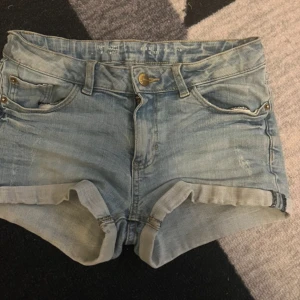 Ljusblå jeansshorts - Snygga ljusblå jeansshorts med upprullad kant. Shortsen har normal passstorlek och har en enkel knapp- och dragkedjestängning framtill. Små fläckar på bakfickan som går bort i tvätten.