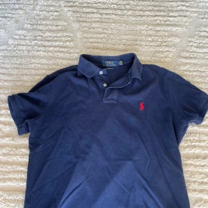 Marinblå pikétröja från Ralph Lauren - Snygg marinblå pikétröja från Polo Ralph Lauren i custom slim fit. Klassisk krage, två knappar och ikoniska röda loggan broderad på bröstet. Perfekt för dig som gillar stilren och sportig look. Storlek M men passar definitivt S