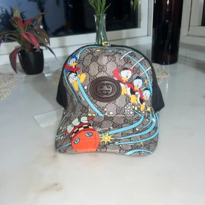 Gucci x Disney keps med Kalle Anka - Snygg keps från Gucci i samarbete med Disney, med Kalle Anka och Knatte, Fnatte och Tjatte på framsidan. Brun bas med GG-monogram, färgglada detaljer och svarta meshpaneler på sidorna. Perfekt statement-piece för dig som gillar unika accessoarer.