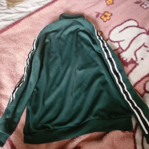 Grön track jacket med ränder från Ath - Mörkgrön track jacket med vita och svarta ränder längs ärmarna och dragkedja framtill. Jackan har hög krage och broderad Ath-logga på bröstet. Perfekt för en sportig och chill stil.