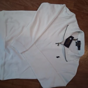 Vit half zip tröja Polo Ralph Lauren - Snygg vit långärmad tröja från Polo Ralph Lauren med half zip-dragkedja och klassisk broderad logga på bröstet. Tröjan är i bomull och har en clean, stilren look som passar perfekt till jeans eller chinos.