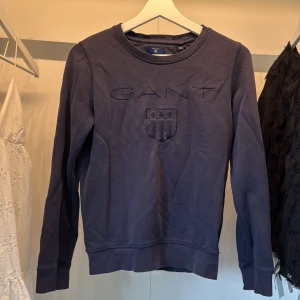 Mörkblå sweatshirt från GANT - Mörkblå sweatshirt från GANT med broderad logga och sköld framtill. Tröjan har rund halsringning, långa ärmar och ribbade muddar vid ärmslut och nederkant. Tillverkad i mjuk bomull för en skön och avslappnad stil.  