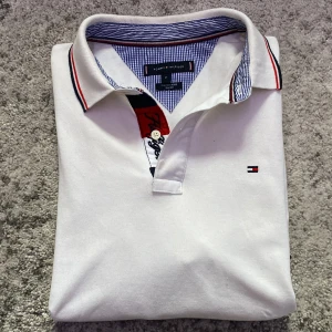 Vit piké från Tommy Hilfiger, slim fit - Vit pikétröja från Tommy Hilfiger i slim fit med krage, broderad logga på bröstet och snygga detaljer i rött och marinblått vid knäppningen. Väldigt sparsamt använd, som ny. Svarar snabbt så hör av er!