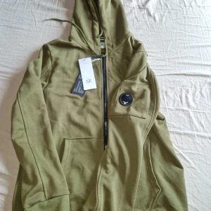 Olivgrön hoodie från C.P. Company - Snygg olivgrön hoodie från C.P. Company med dragkedja och klassisk linsdetalj på ärmen. Tröjan har huva med snörning och praktiska fickor framtill. 