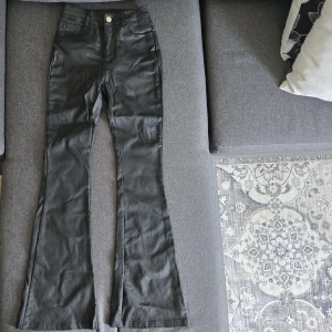 Svarta bootcut byxor med hög midja - Snygga svarta byxor med bootcut och hög midja. Byxorna har klassiska fickor fram och bak, samt bälteshällor. Materialet är mjukt och har en lätt glansig finish som ger en modern look.     Den är i bra skick förutom en liten skada längst ner vid benet (syns på bilden 3).