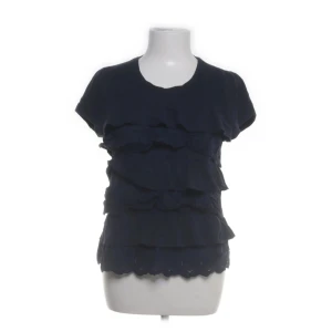 Mörkblå blus med volanger - Supersöt mörkblå blus med flera lager volanger framtill och korta ärmar. Rund halsringning och broderad nederkant ger en extra touch. Perfekt för dig som gillar en feminin och lekfull stil. Liten i storleken!