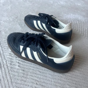 Adidas Samba sneakers mörkblå/vit - Klassiska Adidas Samba sneakers i svart skinn med vita detaljer och bruna gummisulor. Ikoniska tre ränder på sidorna och guldfärgad SAMBA-text. Lågt skaft, snörning och rund tå. Perfekta för dig som gillar retrostil och streetwear. Använda men i gott skick använda fåtal gånger 