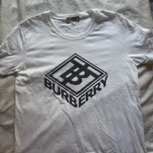 Vit Burberry t-shirt med stor logga - Säljer en clean vit t-shirt från Burberry med stor svart logga och TB-monogram på bröstet. Klassisk rund halsringning och korta ärmar. Tillverkad i mjuk bomull som känns skön mot huden. Perfekt för dig som gillar stilrena designer med lyxig känsla.