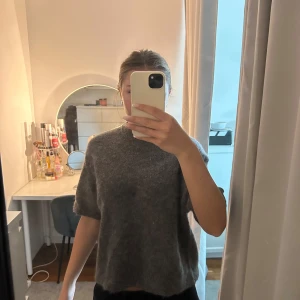 stickad tröja med kort ärm - Mysig stickad tröja med grå/begie/brun nyans på färgen med korta ärmar och rund hals. Tröjan har en loose passform och är perfekt för lager-på-lager-stil under kyliga dagar. Enkel och stilren design som funkar till det mesta.
