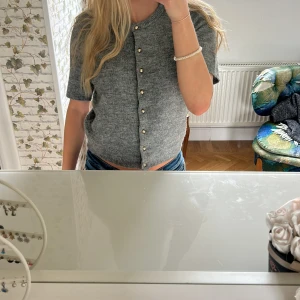 Grå kofta med pärlknappar från H&M - Kortärmad grå kofta från H&M i stickad kvalitet. Tröjan har snygga pärlknappar framtill och en rund halsringning. Perfekt att styla med jeans eller kjol för en chill look.