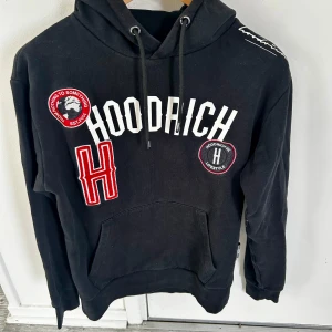 Svart hoodie från Hoodrich med patches - Svart hoodie Hoodrich Pacific med stora vita och röda patches och broderade detaljer framtill. Tröjan har huva med snörning, känguruficka och långa ärmar. Snygg streetstil med tydlig logga och coola emblem. Perfekt för dig som gillar urban vibe. I nederkant på ryggen är det en fläck som inte går bort, se bild. Därav priset. 