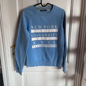 Blå sweatshirt från Gina Tricot - Ljusblå sweatshirt från Gina Tricot med vit text 'New York University N.Y.C.L' framtill. Tröjan har rund hals, långa ärmar och är i mjuk bomullsmix. Perfekt för en chill och avslappnad stil.
