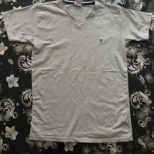Grå  t-shirt Tommy Hilfiger - Snygg grå t-shirt från Tommy Hilfiger med v-ringning och liten broderad logga på bröstet. Klassisk passform och korta ärmar, perfekt till jeans eller shorts. Tillverkad i mjuk bomull för skön känsla hela dagen. OBS!! Jag är Villig att föhandlav priset!!!!