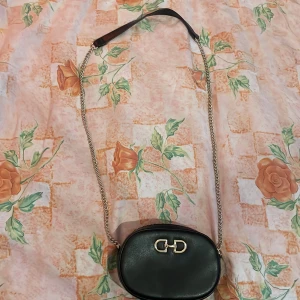 Svart axelväska från H&M med kedja - Snygg oval axelväska från H&M i svart skinnimitation med guldfärgad kedjerem och dekorativt metallspänne framtill. Väskan har dragkedja upptill och en stilren look som passar till många outfits. Perfekt storlek för det viktigaste. Max använd 3 gånger 