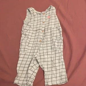 Rutig byxdress från H&M, strl 74 - Supersöt ärmlös byxdress från H&M i vitt med svart rutmönster. Byxdressen har knappar framtill, två fickor på sidorna och en liten rosa hjärt-detalj vid axeln. Perfekt för en chill och lekfull stil till de minsta.