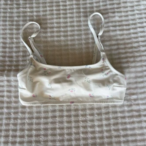 Blommig bikiniöverdel från Lager 157 - Söt bikiniöverdel i ljusbeige från Lager 157 med små blommiga detaljer i rosa och grönt. Modellen har smala, justerbara axelband och en enkel, sportig form. Perfekt för strandhäng och sommarens alla bad.