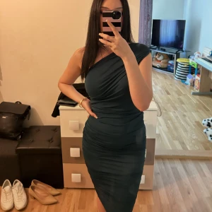 Mörkgrön one shoulder knä-lång klänning - Snygg mörkgrön klänning med one shoulder-design och tight passform. Klänningen är figurnära och har rynkade detaljer längs sidorna som ger en cool look. Perfekt för dig som vill sticka ut med en elegant men modern stil. Passar bra på S och M storlek