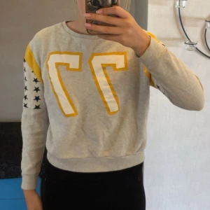 Grå sweatshirt med gula detaljer - Grå sweatshirt från BikBok i storlek XS med gula och vita detaljer samt stora siffran 77 framtill. Ärmarna har svarta stjärnor på vit bakgrund och gula partier vid axlarna. Mjuk och skön, perfekt till chill dagar. Rund hals och ribbade muddar.💛