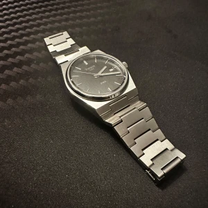 Tissot PRX silver klocka med länk - Säljer en stilren Tissot PRX med svart urtavla och datumvisning. Klockan har en cool, borstad silverfärgad länk i rostfritt stål och levereras med originalask och extra länkbitar. Klassisk design som funkar till det mesta och känns riktigt lyxig på handleden.  Mycket bra skick. 