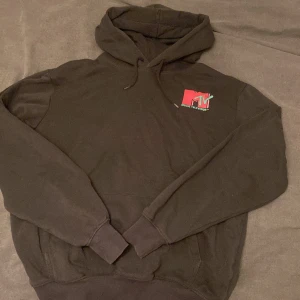 Svart MTV hoodie med tryck - Svart hoodie med klassiskt MTV Music Television-tryck i rosa och mintgrönt på både bröstet och ryggen. Tröjan har huva med snörning och en stor magficka. Perfekt för dig som gillar retrostil och vill sticka ut med ett ikoniskt motiv.