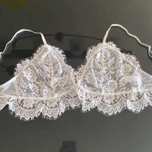 Spetsig bralette med volangkant - Superfin vit bralette i spets med blommigt mönster och volangkant runt hela. Axelbanden är smala och justerbara, och den har hakknäppning bak.