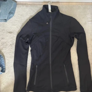 Svart träningsjacka från Lululemon - Slimmad svart träningsjacka från Lululemon med hög krage och hel dragkedja framtill. Jackan har två dragkedjeförsedda fickor och är tillverkad i ett stretchigt syntetmaterial som passar perfekt till träning. Snygga sömdetaljer och diskret logga på ryggen.