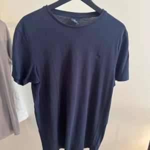 Mörkblå basic t-shirt i bomull från Springfield - En stilren mörkblå t-shirt i mjuk bomull med rund halsringning och korta ärmar. Enkel design med en liten broderad logga på bröstet. Perfekt för dig som gillar en clean och klassisk look.