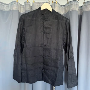 Svart traditionell linneskjorta - Svart skjorta i linne-liknande material med traditionella knytknappar framtill och ståkrage. Skjortan har lång ärm och två fickor framtill. Snygg och unik design som sticker ut från vanliga skjortor. Köpt i Japan