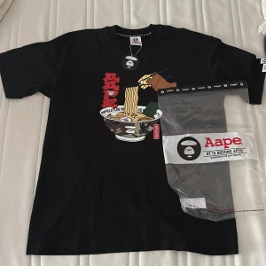 Svart Aape t-shirt med ramen-tryck - Svart t-shirt från Aape by A Bathing Ape med stort färgglatt ramen-tryck på bröstet och röda japanska tecken. Klassisk passform, rund hals och cool streetwear-vibe. Perfekt för dig som gillar unika prints och japansk stil. Köpt i harajuku tokyo juli i år. Helt oanvänd ny pris 900kr