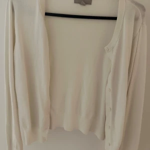 Vit kofta med knappar - En stilren vit kofta med långa ärmar och knappar framtill. Koftan har en klassisk rund halsringning och är tillverkad i ett mjukt, tunt material som känns skönt mot huden. Perfekt att slänga över en topp för en clean look. Aldrig använd