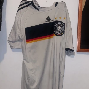 Tysklands landslagströja Adidas - Säljer en officiell Tyskland fotbollströja från Adidas. Tröjan är vit med svart, röd och gul rand över bröstet, broderat landslagsemblem och tre stjärnor. Klassisk rund hals med svarta detaljer och korta ärmar. Tillverkad i lätt och ventilerande polyester.