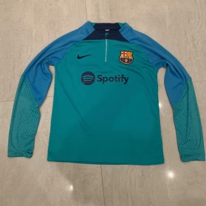 FC Barcelona Nike långärmad träningströja - FC Barcelona långärmad träningströja från Nike i turkos och blått med klubbmärke och Spotify-logga på bröstet. Tröjan har halv dragkedja och är tillverkad i ett lätt, sportigt material som andas. Perfekt för dig som vill visa ditt stöd för Barca på träningen.