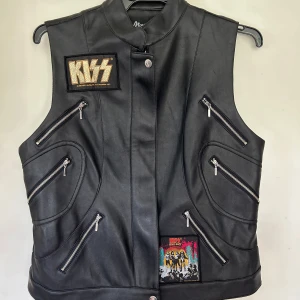 Svart skinnväst med KISS-patchar - Helt ny. Konstskinnväst från Mousance i storlek XXL/48. Tyvärr är det något litet rivmärke på som jag inte vet vad det kommer ifrån eftersom den är helt ny.