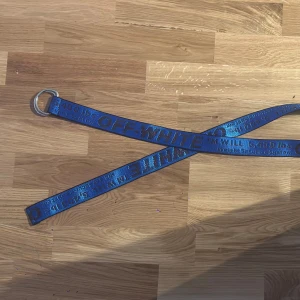 Blå Off-White industribälte - Industribälte från Off-White med tydlig logga och text längs hela bältet. Bältet har en metallring som spänne och är gjort i slitstark syntet. Perfekt accessoar för att lyfta din outfit med streetwear-vibe.