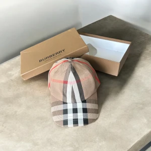 Burberry rutig keps i bomull - Snygg keps från Burberry med klassiskt rutigt mönster i beige, svart, vitt och röda detaljer. Broderad Burberry-logga baktill och justerbar rem. Tillverkad i bomull för en skön och stilren look.