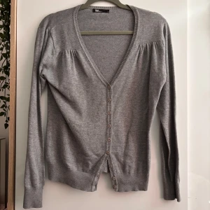 Grå kofta i bomull - En stilren grå kofta från Flash Basic i 100% bomull. Koftan har v-ringning, långa ärmar och knäppning framtill med flera knappar. Extra knapp kommer med ifall den skulle behövas, inga defekter!