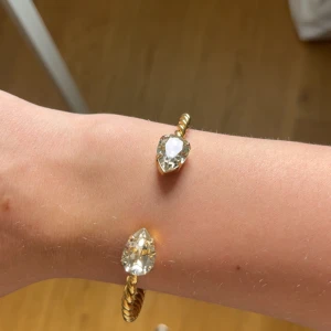 Guldigt armband med stora kristaller - Använd fåtal gånger!! Snyggt armband i guldton med tvinnad design och två stora, droppformade klara kristaller i ändarna. Armbandet är öppet och stela i modellen, vilket gör det lätt att ta på och av. Perfekt statement-piece för att lyfta din outfit.