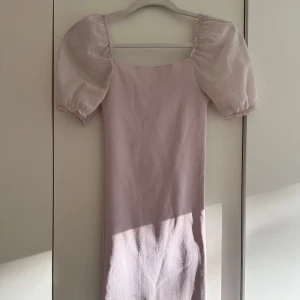 Ljuslila kort puffärmad klänning - Supersöt ljusrosa kortklänning med puffiga, transparenta kortärmar och rak halsringning. Klänningen har en tight passform och är tillverkad i ett mjukt, stretchigt material med släta detaljer. Perfekt för dig som gillar romantisk och trendig stil. Tyvärr har klänningen en liten defekt vid axeln