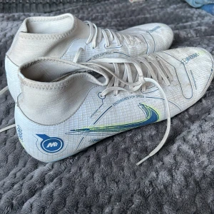 Nike Mercurial vita fotbollsskor - Säljer ett par vita Nike Mercurial fotbollsskor med blå och neongula detaljer. Skorna har snörning, högre skaft och mönstrad syntetisk ovandel för extra grepp. Perfekta för dig som vill sticka ut på planen med stilrena och sportiga skor.
